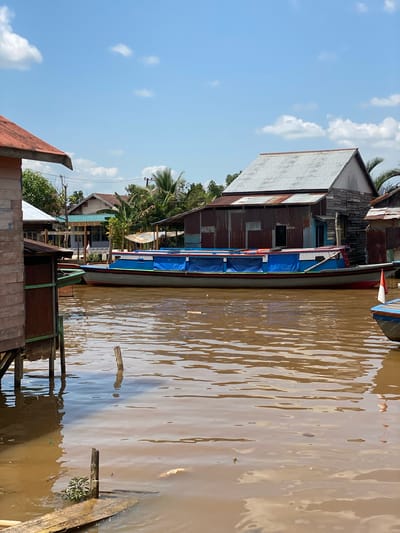 The image depicts a scene in Berangas Timur, Indonesia, characterized by a brown, muddy river flowing through a village. Several wooden houses on stilts are visible along the riverbank, constructed from corrugated metal and weathered wood. A long, flat-bottomed boat, predominantly blue with red and green trim, is moored in the river, covered with blue tarps. A smaller boat with a red and white Indonesian flag is partially visible to the right. The sky above is blue with scattered white clouds. The overall environment suggests a tropical, humid climate.