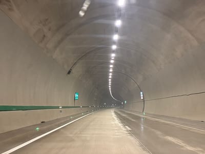 This image shows a view from inside a tunnel, likely on a highway, looking forward. The tunnel is constructed of concrete with a curved ceiling and walls. A line of lights illuminates the tunnel from the ceiling. The road surface is wet, suggesting recent rain or moisture. White lane markings are visible on the road. There are green illuminated signs on the walls, one indicating "135 M" and another with a pictogram of a person and an arrow pointing forward. There are also small green lights embedded in the side of the road. The overall impression is of a modern, functional tunnel infrastructure. There are no people or vehicles visible in the image. The specific location in Achara, Georgia, cannot be determined from the visual information alone.