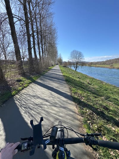 The image presents a first-person perspective from an individual operating a bicycle on a paved path in Hattingen, Germany. The scene depicts a sunny day, with strong shadows cast across the path.

In the immediate foreground, a left hand grips the black rubber handlebar grip of a bicycle. Attached to the handlebars are a black smartphone mount, a digital display unit showing "19.6" (likely speed in km/h) and "69" (likely battery percentage), and a black bicycle headlight. "RACE FACE" branding is visible on the handlebar stem.

The midground is dominated by a dark asphalt path, approximately 3-4 meters wide, extending straight into the distance. To the left of the path, a dense line of tall, bare deciduous trees, devoid of leaves, casts long, intricate shadows onto the path. Underlying these trees are low-lying bushes and ground cover, some with residual greenery. To the right of the path, a gently sloping grassy embankment, with patches of green and brown foliage, descends to a wide river with blue-green water. A solitary, bare deciduous tree stands on the riverbank further down the path.

In the background, the river continues, bordered on its opposite bank by rolling hills and fields with sparse, bare trees and patches of green. The sky is clear and bright blue, with minimal cloud cover visible near the horizon. The overall environment suggests late autumn, winter, or early spring due to the bare trees. No other individuals are clearly visible in the immediate vicinity, though a faint distant figure may be present on the path. This setting is consistent with a riverside cycle path along the Ruhr River in Hattingen.