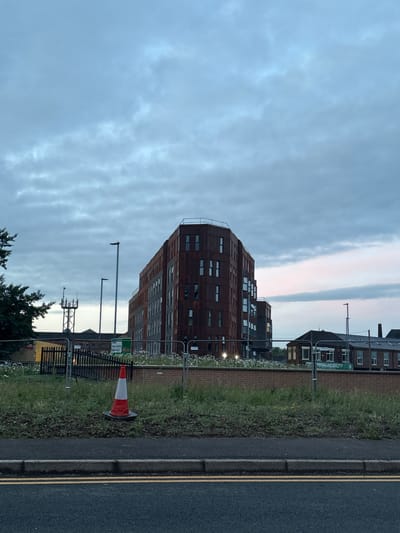 This image captures a quiet urban street scene in Stoke-on-Trent, United Kingdom, likely at dawn or dusk due to the soft, diffused light and subtle pinkish hues in the overcast sky.
The foreground features a tarmac road with a grey curb and prominent double yellow lines, typically indicating no parking. Alongside the road is a grassy verge dotted with weeds and small white flowers. A black metal fence separates this verge from the buildings beyond.
The most striking subject is a modern, multi-story building on the left, characterized by its distinctive dark, possibly weathered or rusted, reddish-brown cladding and an irregular arrangement of numerous rectangular windows. This building appears to be part of a larger structure. To the right, further in the background, an older, traditional brick building with white window frames can be seen behind the fence.
Objects on the verge include several lampposts; two prominent, tall ones in the mid-ground and a smaller, lattice-like structure with antennae further back. A red and white traffic cone lies on its side near the fence.
No people are visible, and no specific activity or event is taking place, contributing to a serene and still atmosphere. The sky is predominantly overcast with varied cloud patterns.
Visible text includes a green sign attached to the fence in front of the older building, clearly displaying "TOTAL MOBILITY" and, beneath it, "Wheelchairs Scooters & Living Aids". Another partially obscured green sign near the traffic cone shows fragments of text like "Stay safe," "Old Com," and "Keep yo...", suggesting a warning or informational notice, possibly related to construction or maintenance.