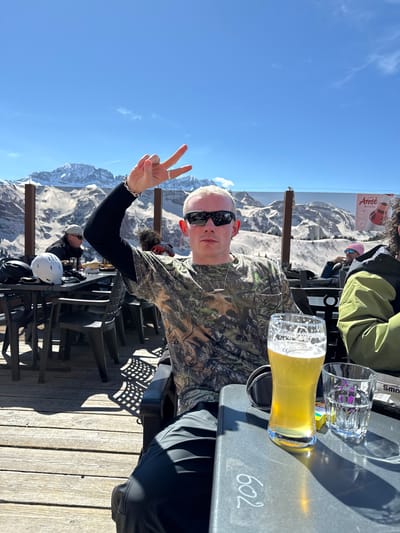 The image captures a man sitting at an outdoor table, making a peace sign with his left hand. He has short, blond hair and is wearing sunglasses and a camouflage shirt. A glass of beer sits on the table in front of him, with other glassware nearby. In the background is a panoramic view of snowy mountains under a clear blue sky. The setting appears to be a terrace or deck, likely a restaurant or bar, with chairs and other patrons visible in the periphery. The table has the number "602" written on it. The man is likely located in Champéry, Switzerland, as evidenced by the mountainous backdrop and signage in the background.