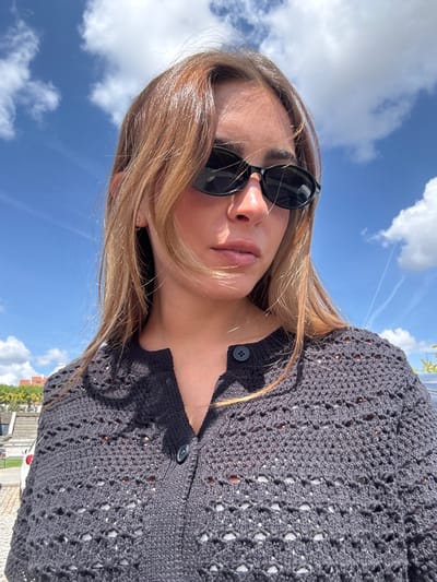 A woman wearing sunglasses and a dark, crocheted cardigan is outdoors in Mogadouro, Portugal. The sky is blue with scattered white clouds. A building with a red roof is visible in the background.