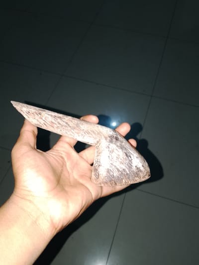 A hand is holding a carved wooden object that resembles a tool. The object is pointed at one end and wider at the other, with visible grain patterns and a rough texture. The background appears to be a tiled floor with shadows cast by the object and hand. The scene is dimly lit. The image was captured in Sendangrejo, Indonesia.