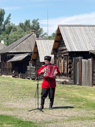 In Shushenskoye, Russia, a man in traditional Cossack attire, including a red tunic, black trousers, and a black fur hat, plays a red accordion. He stands in a grassy area in front of several wooden log houses. A microphone on a stand is positioned in front of him. The sky is blue with some clouds, and trees are visible in the background. The log houses have wooden roofs and appear to be old structures.