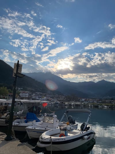 The image shows a view of the harbor in Skala Potamias, Greece, at sunset. The sky is partly cloudy with the sun shining through the clouds, creating a bright glow. Mountains are visible in the background. In the foreground, a boat is docked, and other boats are visible further in the harbor. Buildings line the waterfront and there is a streetlight in the left foreground.
