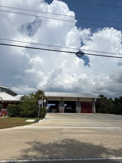 The image shows a modern fire station in Parrish, Florida, United States. The building features a gray metal roof, large red bay doors, and gray siding. Two fire trucks are visible inside one of the bays. In front of the station, there is a red fire hydrant and landscaping. Overhead power lines cross the top of the image, with a wire-hanger-like insulator visible on one of the lines. The sky is filled with dramatic, dark clouds, suggesting an impending storm. The foreground is a concrete paved area, possibly a road or parking lot, with white lines painted on it. The overall scene depicts a functional fire station under a dynamic sky.