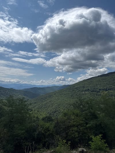 The image depicts a panoramic view of lush, green mountainous terrain under a partly cloudy blue sky. The foreground features dense foliage and some rocks. The middle ground is dominated by rolling hills covered in dense, dark green trees, suggesting a forest. In the background, layers of mountains fade into a hazy blue, indicating distance. Several large, white and gray cumulus clouds are prominent in the sky, casting shadows on the mountains. The scene appears to be a natural landscape, likely a mountainous region. There are no people or animals visible in the image, nor any discernible human structures except for what might be a very small building or structure barely visible on the slope of one of the distant hills, too small to identify with certainty. The overall impression is one of natural beauty and tranquility. The location is described as Manaselska Reka, Bulgaria.