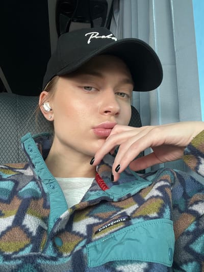 The image depicts a young adult female seated indoors, potentially within a vehicle or a room, in the city of Абухаўскі сельскі Савет, Belarus. The subject is shown from the chest up, facing the camera with a neutral expression. She wears a black baseball cap with a white "Primitive" logo on the front, and a multi-colored Patagonia fleece pullover jacket with teal accents and a visible label. A light grey undershirt is visible beneath the fleece. A white wireless earbud is present in her left ear, along with a small stud earring. Her fingernails are painted black. Her left hand is raised to her face, with fingers touching her chin and lower lip. The background to the left features a dark grey patterned surface, possibly a seatback. To the right, a light blue background is visible, possibly a wall or curtain.