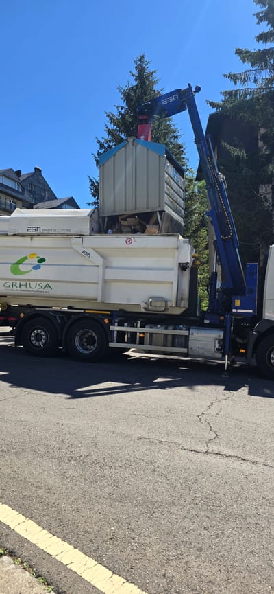A blue crane arm, attached to a white garbage truck, is lifting a large, light-colored debris bin into the back of the truck. The bin appears to be filled with various types of refuse, including wood and cardboard. The truck has "ESA WASTE SOLUTIONS" and a green and yellow logo with the text "GRHUSA Gestión de Residuos Huesca s.a.u." printed on its side. The truck is parked on an asphalt road with a yellow line painted on the edge. In the background, there are green trees and the roof of a building. The sky is clear and blue, indicating a sunny day. The scene appears to be set in a residential or commercial area, possibly during a waste collection service. The image is oriented vertically.