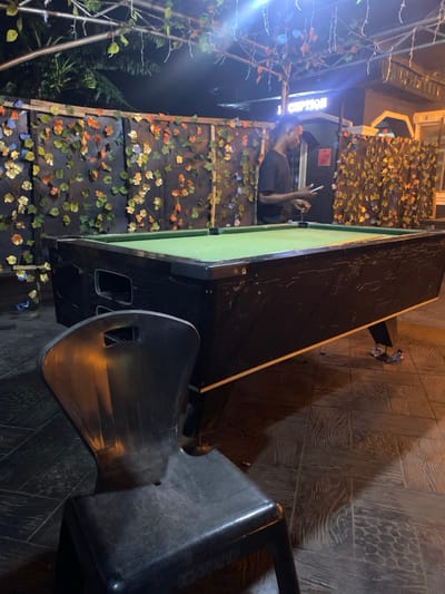 The scene, set in Igbessa, Nigeria, depicts an outdoor or semi-enclosed recreational area at night, illuminated by artificial lighting. An adult male, dark-skinned and wearing a dark short-sleeved shirt, stands to the right of a black pool table with a green felt surface. He holds a white-screened smartphone in his left hand, engaged with the device. The pool table's dark frame exhibits visible wear and scuffs and features a white trim along its bottom edge. No pool balls or cues are present on the table. In the foreground, a dark grey plastic chair is partially visible, facing away from the viewer. The background consists of a dark wall extensively covered with artificial multi-colored leaves (green, orange, yellow, blue). An overhead structure supports hanging strings of decorative lights and more artificial vines. A brightly illuminated sign reading "RECEPTION" is mounted on a building in the upper right background. The ground is covered with dark, textured tiles, and two crumpled pieces of debris are visible near the pool table's base.
