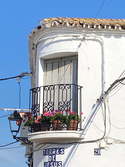 The image depicts a close-up view of the exterior of a white building under a clear blue sky, likely in Cancelada, Spain, as suggested by the street sign. The building features a rounded corner with a small balcony adorned with potted flowering plants, predominantly pink and red. The balcony has a black wrought-iron railing. Above the balcony, a window is covered by a slatted blind. The roofline is characterized by a wavy, terracotta tile edge. Several electrical wires are visible, strung across the building and extending from a black lamppost mounted on the left side. A street sign with blue lettering on white tiles reads "TORRES DE JESUS", and a house number "20" is also visible. The overall impression is of a typical Mediterranean architectural style.