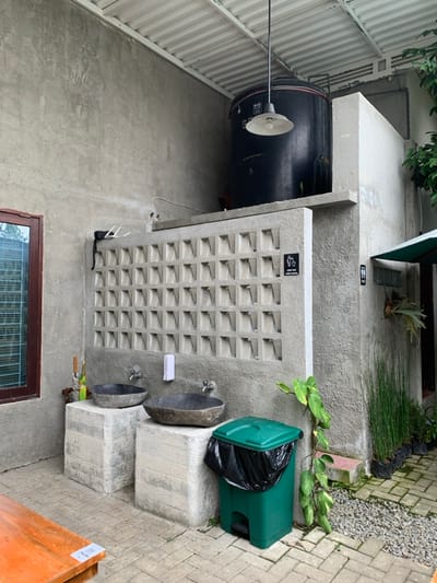 The image depicts a semi-outdoor handwashing station situated in Kota Batu, Indonesia. Two oval, dark grey stone basins, each equipped with a silver-colored faucet, are positioned atop two light grey, rectangular concrete pedestals of varying heights. A white, rectangular soap dispenser is mounted on the textured concrete wall between the basins. Above this, a segment of the wall is composed of light grey concrete vent blocks arranged in a grid. A black sign with a white trash disposal icon is affixed to these blocks. Above the vent block section, a large, black cylindrical water tank rests on a concrete ledge beneath a corrugated metal roof, from which a white, bell-shaped industrial light fixture hangs. To the immediate right, a narrow concrete wall displays a black "Toilet" sign with male and female icons, indicating a nearby restroom entrance. A green market umbrella or awning is partially visible extending from this wall. A large green leafy plant with trailing vines grows against the right wall. A green plastic trash can with a black liner is positioned on the paved ground, which consists of light-colored rectangular pavers. Further right, a row of slender green plants in black plastic bags are arranged on a gravel bed. To the far left, a brown-framed window with horizontal slats is visible on a light grey concrete wall. A portion of a wooden table is present in the lower-left foreground. No individuals are present in the scene.