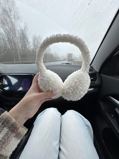 The image, taken from inside a vehicle, shows a hand holding a pair of white fluffy earmuffs against the backdrop of a snow-covered windshield. Outside, barren trees are visible through the windshield. The vehicle's dashboard and entertainment screen are partially visible, along with the driver's side air vents and steering wheel. The occupant of the vehicle is wearing light blue jeans and a plaid jacket. The specific location in Kvarsa, Russia, cannot be determined from the visual data provided.