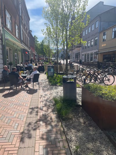 A sidewalk café scene in Kolding, Denmark, is shown on a sunny day. Red brick paving stones comprise the sidewalk, patterned in a herringbone design, with a tactile paving strip. People sit at tables along the sidewalk, including a man in a dark shirt and glasses at the closest table, another man in a white shirt, and several others along the row. Buildings line the street on both sides, with varying architectural styles and colors. A bicycle rack full of bikes is visible behind a planter box filled with green vegetation. The sky is blue with scattered clouds. Shadows from trees are cast on the sidewalk. A gray trash receptacle stands along the street. The name "HESTEN" is visible on a building facade in the background.
