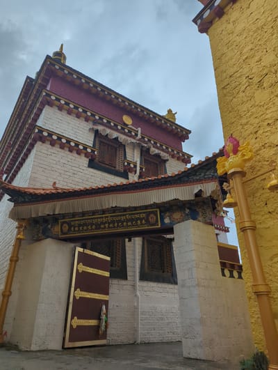 This is a photograph of a building in Shangri-La, China, likely a monastery given the text. The perspective is looking upwards, with a bright yellow textured ceiling in the foreground, possibly a decorative overhang. Below this, the ornate facade of a building is visible. It features red and white brickwork, with decorative elements along the roofline and around the windows. The windows themselves have intricate wooden latticework.

A prominent detail is a vertical yellow banner with gold lettering and intricate designs, which appears to be written in Tibetan and Chinese script, accompanied by English text identifying it as "Shangri-La Ganden Sumtseling Monastery Baden Cashmere Temple". The sky is overcast and a muted grey, suggesting it might be a cloudy day. The overall impression is one of architectural grandeur and cultural significance. The image is vertically oriented.