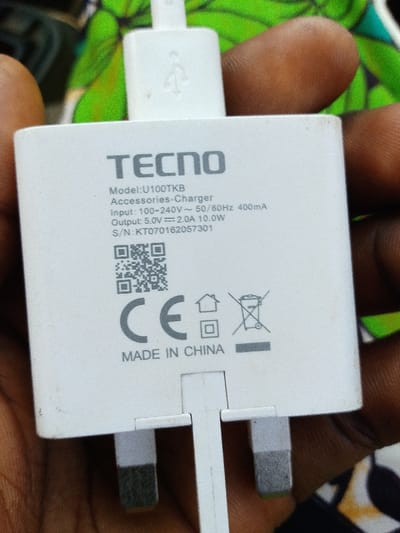 A white TECNO brand power adapter, model U100TKB, is held in a person's dark-skinned left hand. The adapter features a UK-style three-pin plug at the bottom and has a white USB-A cable inserted into its top port, displaying a USB symbol on the connector. Printed specifications on the adapter indicate "Accessories-Charger," "Input: 100-240V ~ 50/60Hz 400mA," "Output: 5.0V === 2.0A 10.0W," and "S/N: KT070162057301." A QR code, CE marking, a house symbol, a double insulated symbol, and a crossed-out wheeled bin symbol are also visible, along with the text "MADE IN CHINA." The background is a blurred, multi-colored fabric, predominantly green and black. This scene is located in the city of Ijara, Nigeria.