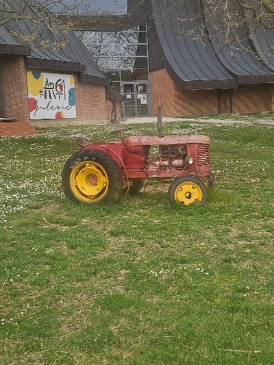 The scene is set outdoors in Avignonet-Lauragais, France, centered on an aged red tractor positioned on a grassy lawn. The tractor exhibits significant weathering, rust, and paint chips, featuring large yellow rear wheels and smaller yellow front wheels. Its engine components are partially exposed, and a vertical exhaust pipe extends from the engine area. The surrounding grass is extensively dotted with small white daisy-like flowers. In the background, a modern building is visible, constructed with brown brick on its lower sections and topped by a distinctive dark, curved or corrugated roof. A central glass-paneled entrance reflects bare tree branches and a visible overcast sky. To the left of the entrance, a large white banner or sign, featuring abstract colorful shapes (yellow, blue, orange, red) and the text "#AR61 Galerie" in black, is affixed to the brick wall. To the right of the entrance, an abstract, dark-colored sculptural figure stands on what appears to be a paved area adjacent to the building. No individuals are present in the depicted environment.