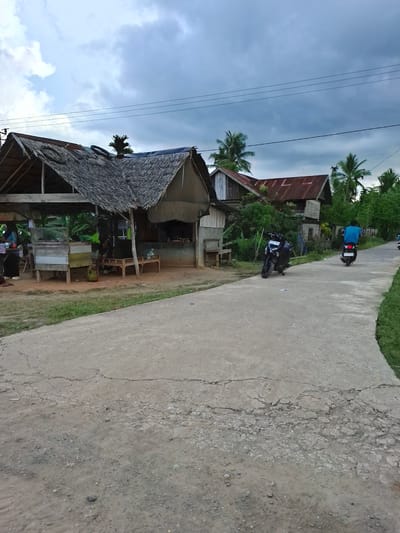 The image shows a rural scene, possibly in Seunebokbaro, Indonesia. A narrow, cracked concrete road stretches through the frame. On the side of the road stands a simple wooden shack with a thatched roof, likely a small shop or residence. Various items are visible within the shack, including tables, chairs, and possibly goods for sale. A person is visible in the shop. Further down the road, another, older house with a rusted corrugated iron roof is partially visible amidst lush vegetation. Two motorbikes are on the road, one parked and one driven by a person wearing a blue shirt. Power lines stretch across the sky. The sky is overcast with clouds. The overall impression is of a simple, rural lifestyle in a tropical climate.