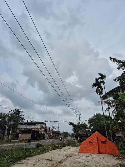 The scene depicts a semi-urban roadside environment in Lagang, Indonesia, under an overcast sky filled with grey and white clouds. In the foreground, an orange, A-frame style tent with two small rectangular vents is pitched on a light brown dirt patch on the right, adjacent to overgrown green vegetation on the left. Multiple black power lines extend diagonally across the upper portion of the frame. In the midground, a dark asphalt road curves slightly to the right into the distance. Along the left side of the road, several weathered buildings with various rooflines and small stalls are visible, accompanied by power poles. On the right side of the road, dense tropical foliage, including tall palm trees and broad-leafed banana plants, partially conceals a small, simple hut with a thatched roof behind the orange tent. Two individuals are operating motorcycles on the road: one in the left lane closer to the foreground and another further down the road in the distance. The overall environment suggests a cloudy day with no direct sunlight.