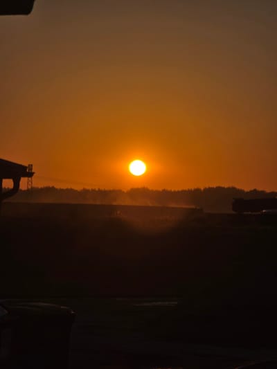This is a vertical, outdoor, medium-distance, full-color photograph of a sunset over a rural landscape. The sun, a bright white orb, is positioned centrally in the frame, slightly above the horizon, casting a warm orange glow across the sky. In the foreground, the silhouettes of dark objects, likely trees and structures, obscure the lower portion of the image. A faint mist or haze hangs low in the distance, partially obscuring a line of trees that forms a dark silhouette against the illuminated sky. On the left side of the frame, a dark, angular structure protrudes from the top edge, possibly a building or roofline. In the far distance, on the right, a dark, large vehicle, possibly a truck, is visible. The overall impression is one of a peaceful, dusky rural scene.