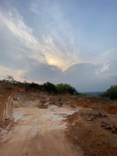 The image captures an open-pit excavation site in Asaga Amangwu, Nigeria, under a partly cloudy sky. The sun, partially obscured by clouds in the upper left-center, casts a bright yellowish light. The ground within the excavation area is characterized by reddish-brown soil and significant deposits of lighter, almost white or cream-colored material. A steep, layered cut of earth displaying both reddish and white strata is prominent on the left. Tire tracks are visible in the lighter-colored earth in the central part of the site. One human figure, an adult in dark clothing, stands in the mid-ground near a pile of white material, facing left. Piles of darker, angular rocks are present on the right side of the excavated zone. Beyond the disturbed earth, a ridge covered with green trees transitions into a hazy, flat landscape extending to the horizon.