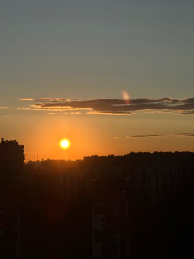 A vertical, eye-level shot captures a sunset over a city. The sun, a bright orange orb, is positioned in the center-left of the frame, casting a warm glow across the sky. Wispy clouds, tinged with orange and pink, stretch across the horizon, some appearing to be illuminated from behind by the sun. A lens flare, a vertical streak of light with rainbow hues, is visible near the top-center of the image. Silhouetted against the vibrant sky is a city skyline, characterized by the dark outlines of buildings and trees. On the far left, a tall, dark building with visible windows stands out. The foreground is dominated by the dark shadows of more buildings and foliage, obscuring many details. The overall atmosphere is serene, with the sunset dominating the visual experience. The image does not contain any people or overt actions.