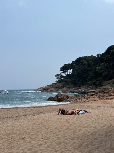 The image depicts a sandy beach in Tamariu, Spain, located at the coordinates 41.918 latitude and 3.208 longitude. The beach is empty except for a couple lying on their backs on a towel in the middle of the sandy beach. The man is lying on his back with his arms outstretched, while the woman is lying on her side facing him, with her arms resting on her stomach. There are a few small rocks in the foreground and a rocky cliff covered in vegetation in the background. The water is a light blue and has small whitecaps. The sky is a light blue and there are no clouds.