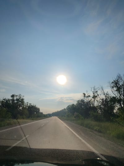 The image presents a forward-facing view from inside a vehicle traveling along a straight, two-lane asphalt road. A brilliant, intense sun is centrally positioned in the upper portion of the clear blue sky, which features scattered, wispy white clouds.
Dense green trees and bushes line both sides of the road, forming a natural corridor. Some trees on the right side appear to have bare branches, contrasting with the lush foliage. The road itself is marked with a solid white line on the right shoulder and a broken white line dividing the two lanes. Further in the distance, the faint outline of another vehicle can be seen on the road ahead.
In the foreground, reflections and shadows from the vehicle's interior are visible on the windshield, including a distinct patterned texture on the left side, possibly from a dashboard mat or object. Partially obscured by the trees on the right side of the road is a yellow, triangular road sign with a red border, likely indicating a warning.
The scene depicts a journey under bright daylight conditions, suggesting it is mid-day or late afternoon given the sun's high position. The weather is clear and fair. The setting is a rural or semi-rural highway in Sofiivka, Ukraine. No explicit text is visible within the image.
