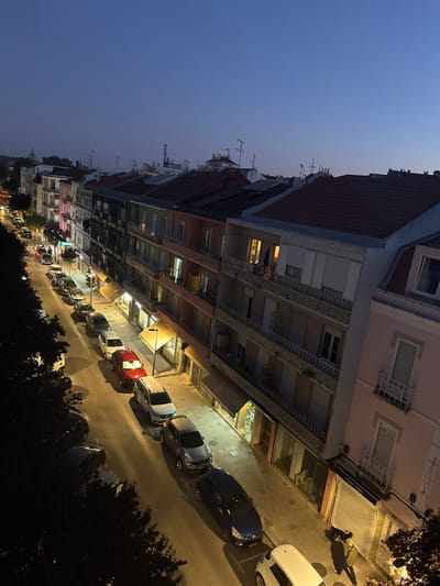 An elevated, oblique, wide-angle shot captures a street scene at dusk in Algés, Portugal. The twilight sky is a deep blue, transitioning to lighter shades near the horizon. Buildings line both sides of the street, featuring multiple stories with apartments above ground-floor commercial spaces. Red-tiled roofs are visible, and many buildings have balconies with decorative railings.

The street is paved and lined with parked cars. From the foreground, moving up the street, are a white hatchback, a dark blue sedan, a silver SUV, a white crossover, a red SUV, a silver sedan, a darker sedan, and several other vehicles further up the street. Streetlights cast pools of yellow light on the road and sidewalks.

On the right side of the street, several storefronts are visible, some with closed metal roll-down shutters, others with awnings. One storefront has a sign reading "Flores Algés." A person is walking on the sidewalk, casting a long shadow. Trees with dark foliage flank the left side of the street, obscuring some of the buildings. The overall impression is a quiet residential and commercial street in a European town as evening settles in.