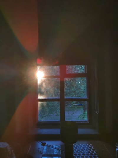 The image depicts an interior scene looking out a window. The sun is setting or rising, casting strong light rays and colorful refractions across the left wall and onto the window panes. Through the window, lush green foliage and a hint of a brick wall are visible, suggesting an outdoor environment with trees.
In the foreground, on a wooden floor, are several objects. To the left, a portion of a woven object is visible. In the center, a dark, cylindrical object stands on a patterned surface, possibly a rug or a tiled floor. The floor itself appears wet or reflective in places, with dark lines or objects present. On the right side of the room, a thin chain hangs vertically against a dark wall.
The overall lighting is dramatic due to the sun's intensity and the resulting lens flares and light scatter. The scene appears to be set in a room with dark walls and a wooden floor.
While the exact location within Лынтупскі сельскі Савет, Belarus cannot be definitively determined from the visual information alone, the presence of dense foliage and a brick wall could be consistent with residential or rural areas within that region of Belarus. The style of the window, with its multiple panes, is also common in older European architecture.