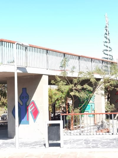 This image depicts an outdoor scene in Camarones, Chile, under a clear, bright blue sky with direct sunlight creating prominent shadows. The central feature is a concrete elevated pedestrian walkway or bridge, supported by visible concrete pillars. This walkway is equipped with a railing system composed of vertical silver-colored metal bars topped by a reddish-brown handrail.

At ground level, the area beneath and in front of the walkway is paved with irregularly shaped dark and light grey stones. A white freestanding structure with a black rectangular panel, possibly a waste bin or signage, is positioned on this paved area near the left concrete pillar.

Graffiti is present on the concrete support structures. On the left pillar, a stylized blue figure with two small circular elements, resembling a head and torso, is depicted alongside a red geometric shape with internal lines, both outlined in light blue. Further right, on another concrete wall supporting the walkway, a teal and green geometric pattern is visible.

Several trees are present throughout the scene. Small leafy plants grow on the elevated walkway structure near the railing. Larger trees with green foliage are visible at ground level, both in the foreground and behind the main structures. To the right, behind a lower metal railing, faint outlines of outdoor exercise equipment can be discerned among the trees.

In the far background, against the clear sky, a tall, slender white metallic structure, resembling a stylized Christmas tree adorned with a spiraling green garland and topped with a star, is prominent. No individuals are present in the image, and no actions or interactions are depicted.