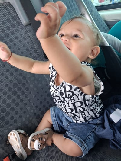 A baby sits in a car seat on what appears to be a bus or passenger van, with gray patterned fabric covering the seats. The baby, with light skin and dark eyes, is wearing a black and white patterned tank top with the word "BABY" repeated in large letters, and denim shorts. The baby's arms are raised, with the right arm extended upwards and the left arm bent and resting on the chest. The baby is also wearing brown sandals, one of which has a cat or tiger face design. A red string is tied around the baby's left wrist. In the background, a stroller is visible, along with a window that shows a blurred outdoor scene, suggesting movement. The image appears to have been taken inside a vehicle. The location of Kukorevo, Bulgaria is not depicted in the image.