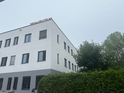 The image shows a modern, white building with a gray lower section in Teltow, Germany. Red lettering on the top of the building spells out "REHA-ZENTRUM". The building has multiple windows on its upper floors and a row of windows on the lower floor. A thick, green hedge runs along the front of the building, and trees are visible to the right. The sky is overcast. The scene appears to be an exterior view of a rehabilitation center.