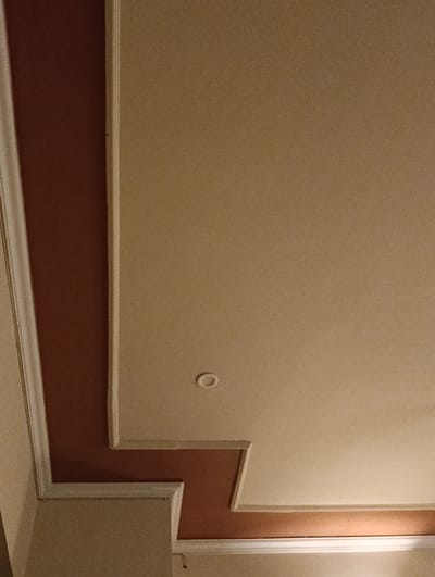 The image shows a section of an interior ceiling with a recessed design. The ceiling features two distinct colors: a lighter beige and a darker reddish-brown. A decorative molding or trim separates these colors and also frames the recessed areas. A small, circular, white fixture, possibly a light or vent, is visible on the beige portion of the ceiling. The lighting in the image is warm, casting a soft glow on the surfaces. The scene appears to be indoors, likely in a residential or commercial building. There is no information about the city of Keer, Pakistan, or any specific location within it that can be determined from this image.