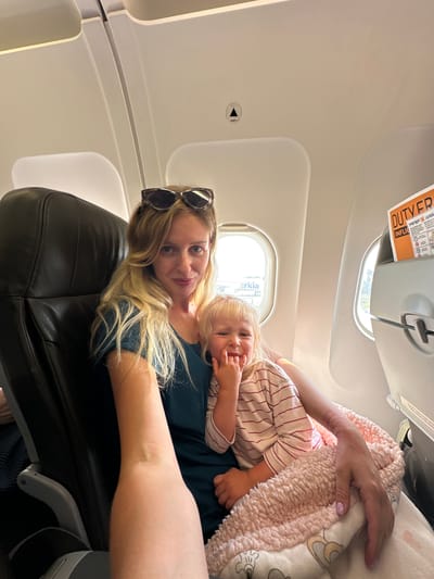 A woman and a young child are seated on an airplane. The woman, with blonde hair and sunglasses perched on her head, is wearing a dark blue t-shirt. She is holding the child, who has blonde hair and is wearing a pink and white striped shirt. The child is looking at the camera with a playful expression. They are both covered by a pink, fuzzy blanket. The environment appears to be a standard airplane cabin, with seats, windows, and overhead compartments visible. A "DUTY FREE" sign is attached to an overhead compartment. The image was taken in Yehud-Monosson, Israel.