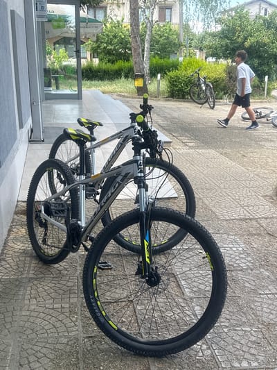 The image depicts a sidewalk in Ovoshtnik, Bulgaria, with several bicycles parked near a building entrance. In the foreground, two mountain bikes are positioned side-by-side. The bike on the right has a black frame with yellow accents on the seat and handlebars, and its front wheel is prominently featured, showing the "GALAXY" branding on the tire. The bike on the left is partially obscured but appears similar in style. Further back, a young boy wearing a light blue t-shirt and black shorts walks away from the viewer towards the right side of the image. He is on a paved area with a pattern of square tiles. Behind him, another bicycle is parked near a bush. The environment suggests a public space, possibly in front of a shop or establishment, with trees and buildings visible in the background.