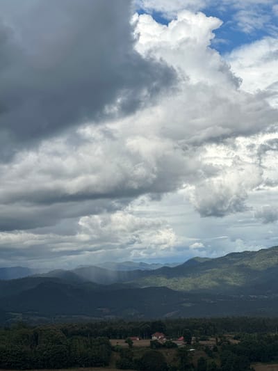 An elevated panoramic view captures a mountainous landscape in Ban Mai Saha Sampan, Thailand. The sky is dominated by a complex cloud system, with dark grey, heavy cloud cover occupying the left portion of the frame, transitioning to brighter white cumulus clouds with visible patches of blue sky towards the upper right. Multiple layers of verdant, tree-covered mountains extend into the background, exhibiting atmospheric perspective. In the mid-distance, distinct shafts of rain are visible descending from the clouds onto a section of the mountains. The immediate foreground, particularly the lower right, features a small cluster of structures, including at least two buildings with reddish-brown tiled roofs, situated amidst cleared or cultivated land and surrounded by dense green trees. No human figures are present.