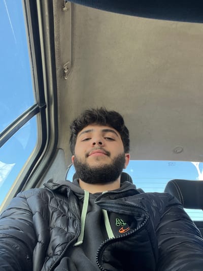 A young adult male, with dark curly hair and a dark beard, is seated inside a vehicle, looking directly at the camera. He wears a dark, possibly black, puffer jacket over a dark hooded sweatshirt with a light green drawstring. On the right chest area of the puffer jacket, a dual-colored emblem, featuring a green and an orange stylized shoe or boot, is visible. The vehicle's interior includes a light grey, upholstered ceiling with visible fasteners, and dark framing around the windows. Through the vehicle's windows, a clear blue sky with minimal white cloud formations and faint contrails is visible, indicating an outdoor, daytime setting. The exact location within the city of Al-Kiswa, Syria, is not visually discernible from the image itself.