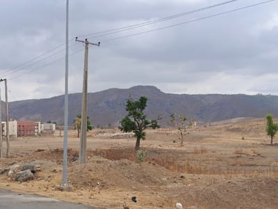 The image depicts an expansive, arid landscape in Rafin Pa, Nigeria, under an overcast sky. In the foreground, a dark gray paved surface is visible on the bottom left, leading to an undeveloped terrain characterized by light brown soil, dry vegetation, and scattered piles of dirt and concrete debris. Three utility poles with overhead power lines traverse the upper midground; one tall metal pole stands centrally, flanked by two shorter wooden poles. Several sparse trees with minimal green foliage are distributed across the dry field, with one prominent green-leafed tree slightly left of center. In the far midground, to the left, a cluster of multi-story buildings, featuring brick-red and beige facades, is visible. Further right, a light-colored water tower or similar structure stands against the distant mountains. The background is dominated by a range of rugged, brown-gray mountains with sparse vegetation. No individuals, specific actions, or interactions are observable.