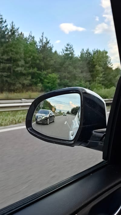 The image shows a reflection in a car's side mirror, depicting a dark-colored sedan driving on a highway, seen from the perspective of another vehicle. The reflection also shows a white car to the right, as well as a distant car. The background visible in the mirror includes a roadside with vegetation and a cloudy sky. The foreground shows the interior of the car from which the photo is taken, including the side mirror assembly and a portion of the car door. The overall scene suggests a journey on a road surrounded by a forested area under a partly cloudy sky. The location of Dolno Varshilo, Bulgaria, is mentioned, but there is no specific visual information within the image to confirm or deny this location.