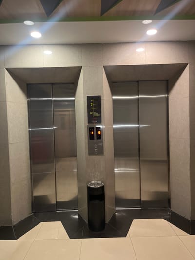 An interior shot shows two identical elevators side-by-side in a hotel lobby. The elevators are clad in stainless steel and have visible seams. To the right of the right elevator, a control panel is mounted on the wall. This panel features a digital display showing the number '5' and a series of buttons, including two circular ones below the display. Above the control panel, a directory lists the floors: '4 guest room', '3 guest room', '2 guest room', and 'GF lobby, restaurant, max health club, swimming pool'. The elevator doors are illuminated by overhead recessed lighting, which is also reflected on the polished surfaces of the elevator doors and the tiled walls. The floor in front of the elevators is tiled in a light cream color, with a darker tiled area forming a geometric pattern around the entrance. A black cylindrical trash bin is situated to the left of the right elevator. The ceiling features a decorative pattern of alternating dark and light wood-like panels, separated by green accents. There are no people visible in the image. The scene appears to be located in Dumai City, Indonesia.
