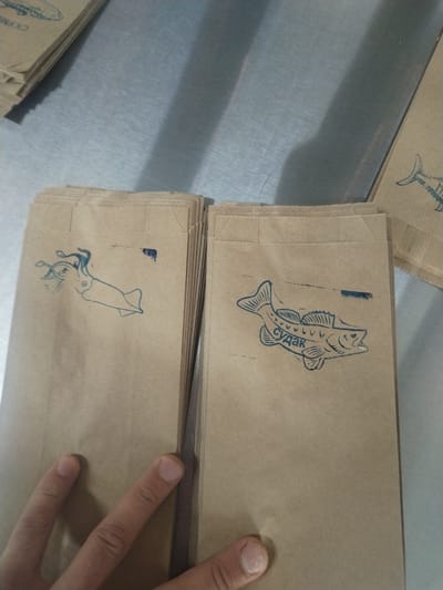 The image shows two stacks of brown paper bags on a metallic surface. The stack on the left has an image of a squid printed on it in blue ink. The stack on the right has an image of a fish, specifically a zander, printed on it, also in blue ink, with the word "судак" (sudak) printed in Cyrillic letters on the fish's body. A hand partially covers the bottom of both stacks. The scene is located in Pionersky, Russia.