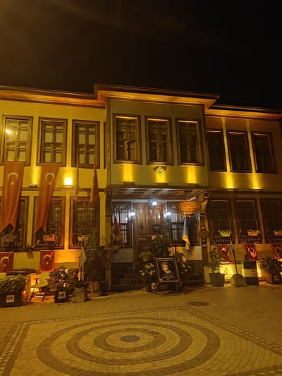 An exterior shot at night shows a historic, two-story building, likely a hotel or cultural center, in the city of Osmangazi, Turkey. The facade of the building is painted a pale yellow, with brown wooden window frames and trim. Warm, yellow lights illuminate the building's architectural details and highlight the windows. Several Turkish flags, orange with white crescents and stars, are draped from the upper story windows. Below the windows, there are flower boxes and potted plants. In front of the building, there is a paved walkway with a circular, brick pattern. To the right of the entrance, a framed portrait of a woman is displayed on an easel, with a small plaque below it. Several large, industrial barrels are also visible near the entrance. The overall impression is one of a well-maintained, traditional building with a festive atmosphere.