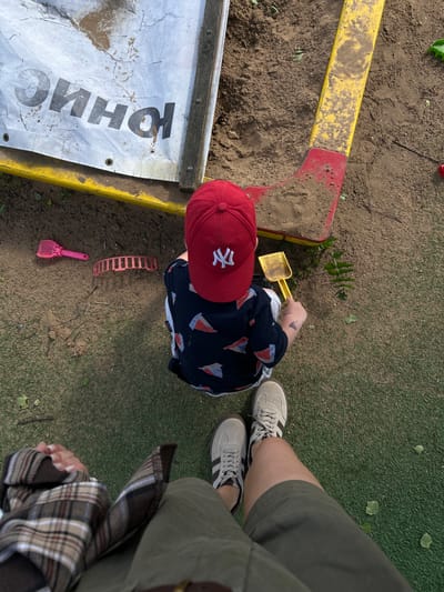 The image presents an overhead view of an outdoor scene. A child wearing a red baseball cap with a white logo, a dark shirt patterned with sailboats, and light-colored pants is playing in a sandpit. The child is holding a yellow plastic shovel. Around the sandpit are various toys, including a pink shovel and a red sand mold. The sandpit has a yellow and red rim on one side and a white and yellow rim on another side with Russian text on it. A portion of an adult's legs and shoes are visible on the right side of the image. The ground surrounding the sandpit is covered with artificial grass and some dirt. The location is outdoors, possibly in Борщевка Малая, Russia.