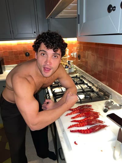 The image shows a muscular, shirtless man with curly hair flexing his arms in a kitchen. He has an open mouth and a playful expression. Behind him, on a white countertop, are several large, bright red prawns or langoustines. A knife and a smartphone are also visible on the counter. The kitchen has a tiled backsplash in a reddish-orange hue. Cabinets are gray. The image is rotated to the left. Based on the metadata, the location is likely Gondarém, Portugal.
