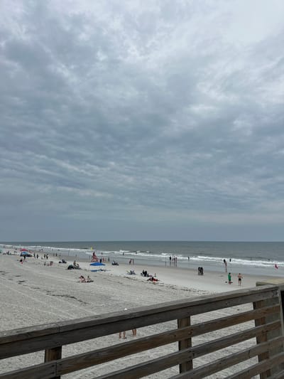 The image shows a beach scene in Jacksonville Beach, United States of America, taken from an elevated perspective near a wooden fence or walkway. The beach is crowded with people engaged in various activities like swimming, sunbathing, and walking along the shore. The sky is overcast with clouds. The horizon line is slightly blurry, possibly due to atmospheric conditions. The wooden fence runs along the right edge of the image, partly obscuring the view of the beach.