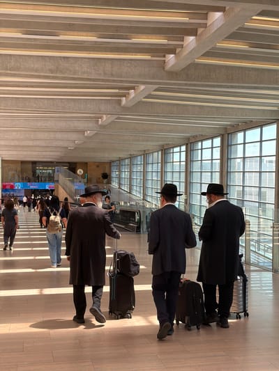In Tzafria, Israel, three men in dark suits and hats walk down a brightly lit hallway within what appears to be an airport terminal. The men are walking away from the viewer, with their backs to the camera. They are each pulling rolling luggage. The hallway features a modern ceiling with exposed beams and integrated lighting, and large windows line the right side of the hallway, offering a view of the exterior. Other people are visible in the background, walking in the opposite direction and towards escalators. The floor is made of polished tiles that reflect the sunlight streaming in from the windows, creating distinct shadow patterns. Digital displays are visible in the distance on the left side of the hallway.