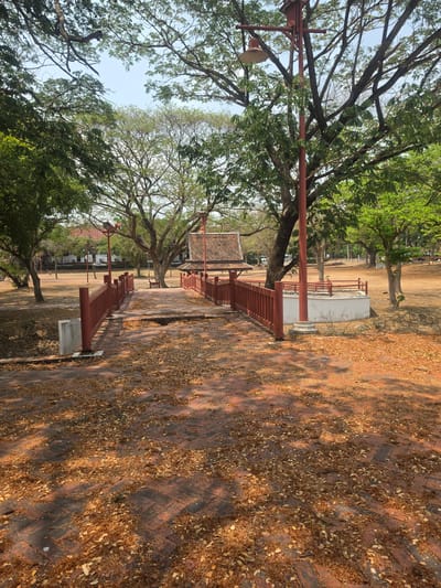This is an outdoor scene in the Historic City of Ayutthaya, Thailand, featuring a brick-paved area covered in fallen leaves. A red-painted railing runs along the side of the walkway, with trees casting shadows across the path. A lamp post with a light fixture is visible on the left, and a traditional Thai-style building with a tiled roof is seen in the background, further indicating the location. The scene appears to be a tranquil, historical site, possibly a park or garden within the city.