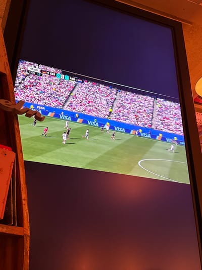 The image shows a television screen displaying a soccer match. The score displayed on the screen is Germany 0, Paraguay 1, with 45:00 minutes played and an additional 1:05 minutes. The match is being played in a stadium with a large crowd in the stands. Several players in white and red uniforms are visible on the green field. The FIFA and VISA logos are also visible on advertisements around the field. The scene appears to be indoors, with part of a wooden shelf visible in the foreground on the left side of the image. There is no visible information in the image to indicate the location in Langeoog, Germany.