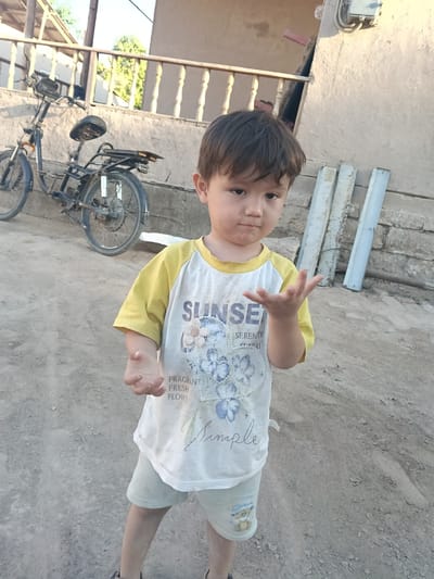 A young boy stands in front of a tan wall in Altinkul, Uzbekistan, holding his right hand up with fingers splayed. He wears a yellow and white t-shirt with graphics and light-colored shorts. Behind him, a dark-colored bicycle is parked against the wall. The ground is dirt and gravel.
